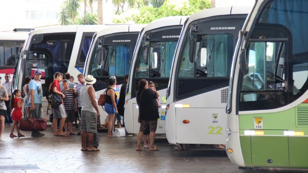 A foto mostra o setor de embarque de ônibus da Estação Rodoviária de Porto Alegre com seis ônibus estacionados. Há filas de passageiros em dois deles. São adultos e crianças com bagagens.