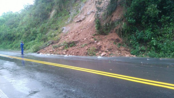 A foto mostra parte de uma encosta desmoronada sobre parte de uma estrada pavimentada, em trecho reto. Um funcionário do Daer, com uniforme azul, observa o monte de terra desmoronado.
