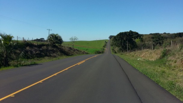 A foto mostra uma estrada em trecho reto, com pavimento e sinalização novos. Ela segue ao horizonte, com vegetação rasteira e árvores às margens.