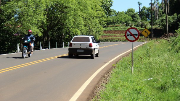 A foto mostra o trecho asfaltado e sinalizado (com pintura e placas de trânsito) da rodovia Johann Martin Kroth, em Venâncio Aires. Um carro e uma moto trafegam em sentidos opostos. Ao fundo, vegetação e propriedades rurais.