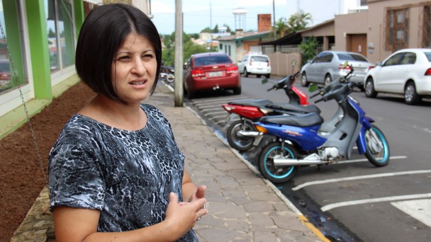 Na foto, a vendedora Vandréia aparece gesticulando, falando com outra pessoa não mostrada na imagem. Ao fundo, uma rua de Taquari, com carros e motocicletas estacionadas.