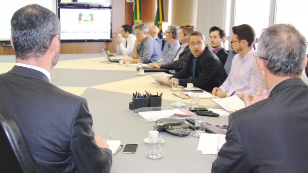 A foto mostra uma reunião ao redor de uma grande mesa. Em destaque, o diretor-geral do Daer, Rogério Uberti, acompanhado de outros diretores, faz uma explanação. Ao fundo, um monitor com o brasão do Estado.