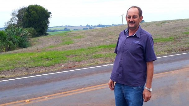 A foto destaca o agricultor Itomar Minuzzi, que está de pé em frente à RSC-163. Ao fundo, campo e vegetação.