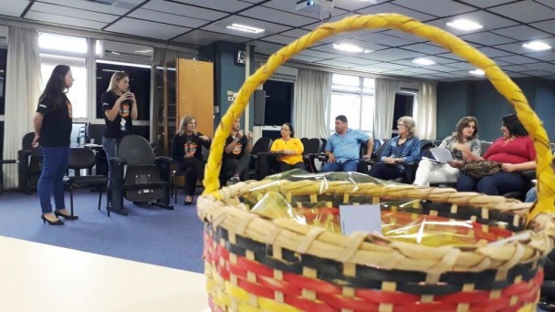 A foto mostra um grupo de professores sentados em um auditório, enquanto assistem a aulas de educadoras do Detran, de pé. Em primeiro plano, uma cesta de palha feita por índios.