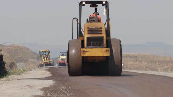 Na foto, uma patrola circula por um trecho em restauração de uma rodovia pavimentada. Ao fundo outras máquinas atuam na obra.