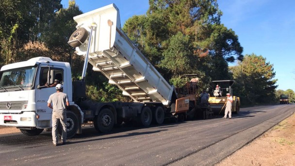 Na foto, um caminhão descarrega material asfáltico na RS-431, para as obras de recuperação da rodovia. Funcionários trabalham na ação, sendo que um deles dirige um rolo compressor logo atrás.