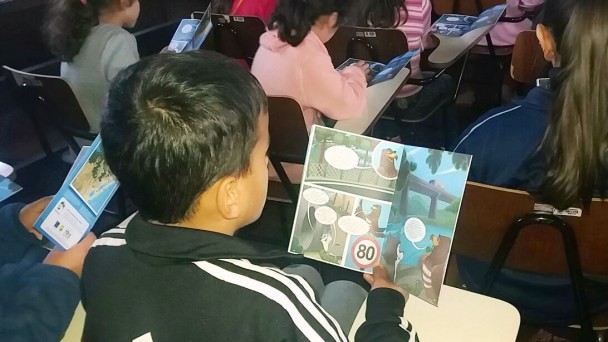 A foto mostra uma sala de aula, com os alunos sentados e de costas, lendo a história em quadrinhos da cartilha "Graxa e sua Turma", produzida pelo Daer.