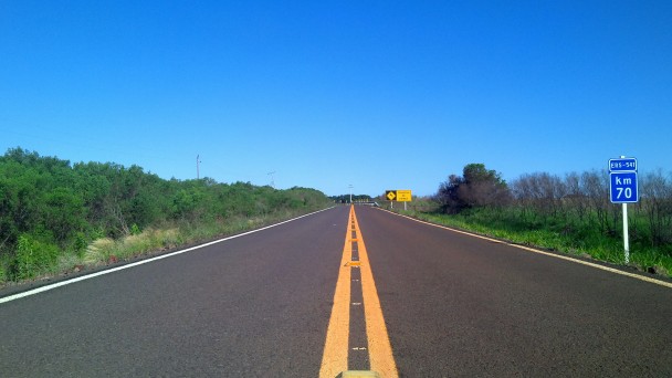 A imagem mostra a rodovia pavimentada e sinalizada com pintura do eixo central em amarelo e das laterais em branco. Na figura, também estão placas indicativas do quilômetros onde foi tirada a imagem. 