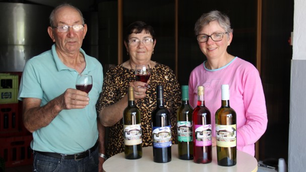Na foto, três comerciantes, sendo um casal e a filha, posam atrás de uma mesa com garrafas de vinho, dentro de uma cantina. Eles seguram taças de vinho.