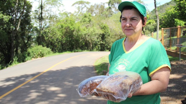 Na foto, a comerciante Veronica Montemezzo posa ao lado da rodovia asfaltada Caminho das Pipas. Ela segura um pão e uma cuca, produzidos por ela. Está uniformizada com a camisa e boné da agroindústria que administra.