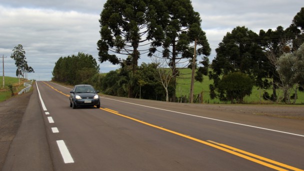 A foto mostra uma rodovia com asfalto e sinalização renovada. Um carro circula por ela. Ao fundo, árvores e campo verde.
