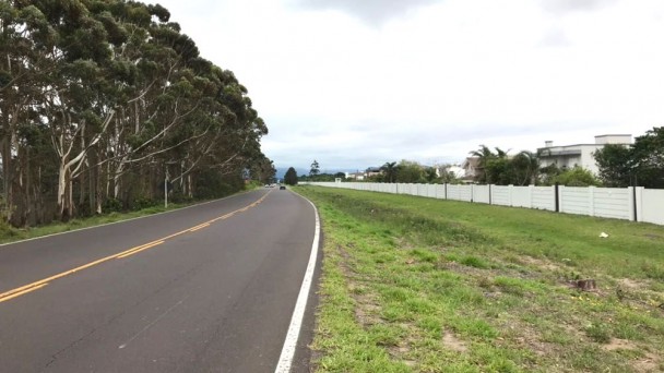 A foto destaca uma área de gramado às margens da Estrada do Mar, em Xangr-lá. Ao fundo, à direita da rodovia, aparece o muro de um condomínio fechado. À esquerda, uma área com árvores.