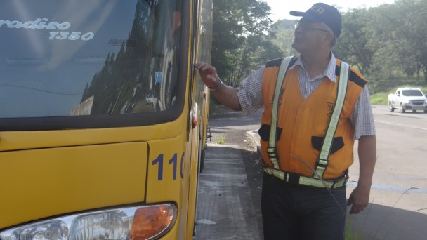 A foto mostra um fiscal do Daer, com colete laranja e boné azul, andando ao lado de um ônibus estacionado às margens de uma rodovia.