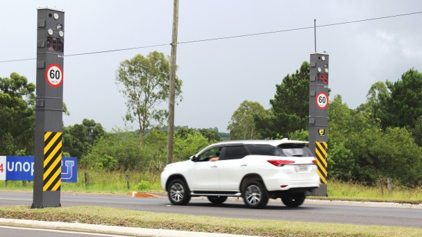 A foto mostra uma caminhonete branca passando por um trecho de rodovias monitorado por duas lombadas eletrônicas.