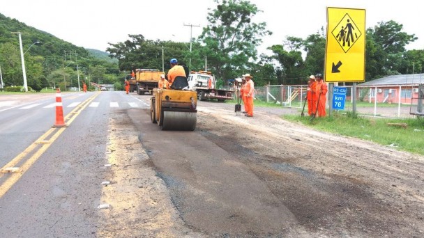A foto mostra um grupo de funcionários do Daer uniformizados com macacões laranja, em obras de manutenção de uma rodovia em frente a uma escola. Um dos operários dirige um rolo compressor.