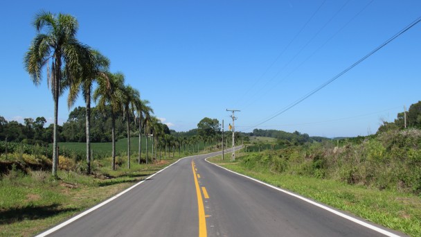 A foto mostra a rodovia de acesso ao município de São Valentim. O asfalto e a sinalização estão em boas condições. Palmeiras contornam a estrada e o céu azul completa a paisagem.