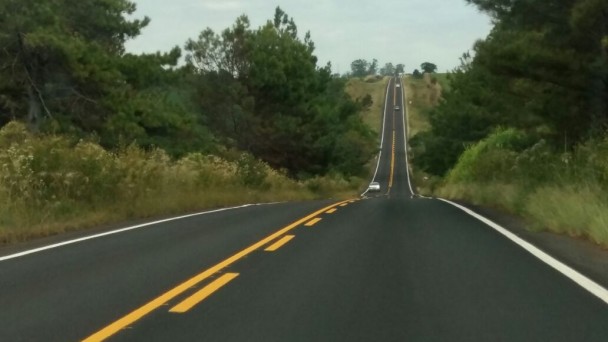 A foto mostra uma rodovia com pista íntegra, sem imperfeições. Ela apresenta pintura marcada nos eixos da pista e no meio dela. Um carro branco transita ao longe.