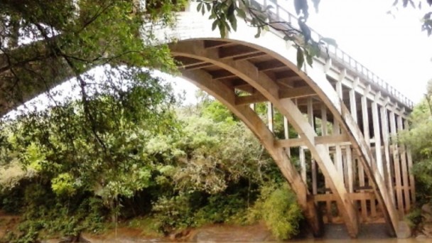 A foto apresenta uma vista da ponte sobre o Rio dos Sinos, na ERS-020, no nível das fundações. Há árvores e vegetação densa às margens do rio.