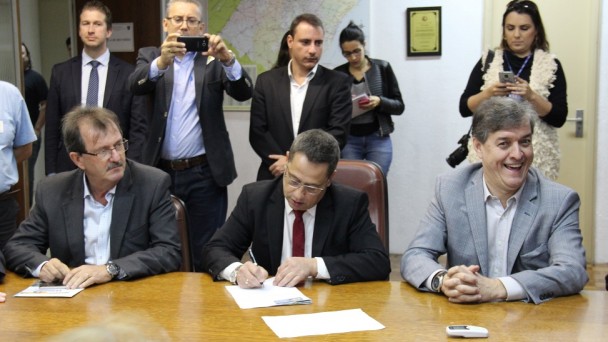 A foto mostra o diretor-geral do Daer, Rogério Uberti, sentado numa mesa de reuniões e com uma caneta na mão durante a assinatura do contrato de pavimentação da ERS-528. Ao lado dele, estão duas autoridades políticas.
