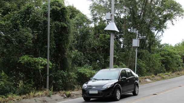 A foto mostra o pardal instalado na descida da curva da morte, no quilômetro 46 da ERS-122. Um carro preto está trafegando na estrada e passa em frente ao dispositivo.Ao fundo, a vegetação às margens da rodovia.
