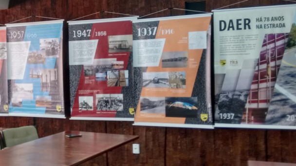 A imagem mostra cinco painéis com fotos antigas do Daer, cada um referente a uma década de história. Eles estão juntos a uma parede de madeira. À frente, no canto esquerdo da foto, uma mesa com cadeiras