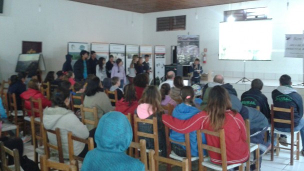 Cerca de vinte adolescentes sentados em cadeiras de madeira, dentro de um ginásio, assiste, a uma apresentação em um telão. Ao fundo, um grupo de jovens de pé, em frente aos banners da exposição do Daer sobre a Rota do Sol.