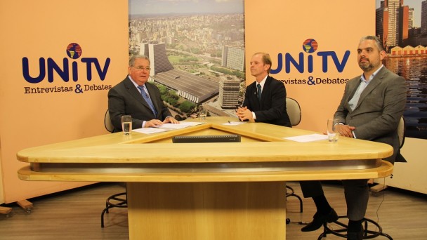 Em um estúdio de TV, sentados ao redor de uma bancada em formato triangular, o jornalista Carlos Alberto Carvalho entrevista os diretores do Daer Ricardo Nuñez e Saul Sastre.