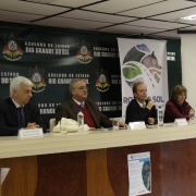 A foto mostra uma mesa com autoridades, entre as quais o diretor-geral do Daer, que fala ao microfone. Ao fundo, um telão projeta foto do diretor com o currículo dele.