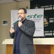 A foto mostra um homem, de terno, barba e óculos, com um microfone, ministrando uma palestra. Ao fundo, uma mesa, um painel com o logotipo da empresa STE e uma tapadeira com vários brasões do Rio Grande do Sul.