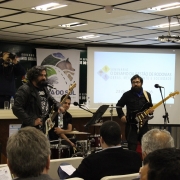 A foto mostra um grupo musical, com guitarras, baixo, e bateria, se apresentando em frente a pessoas sentadas em cadeiras em um auditório. Ao fundo, um telão com o logotipo do seminário do Daer e um painel com o logotipo da Rota do Sol.