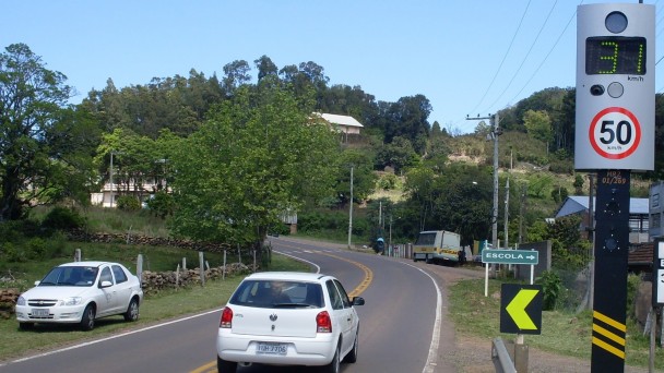 À direita da foto, uma lombada eletrônica indica a velocidade de 31 quilômetros por hora no visor, enquanto, na rodovia, um carro branco passa ao lado. Um outro carro branco está estacionado às margens no sentido contrário. Ao fundo, casas e vegetação.