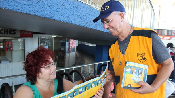 A foto mostra um fiscal do Daer, com boné azul e colete laranja identificado com o brasão da autarquia, entregando um fôlder a uma mulher que está sentada em um dos bancos do setor de embarque da rodoviária de Porto Alegre.