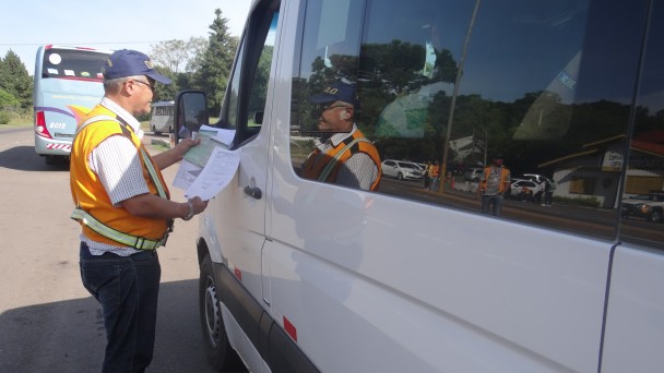 A foto mostra um fiscal do Daer, uniformizado com boné azul e colete laranja, analisando documentos ao lado de uma van de transporte de fretamento.