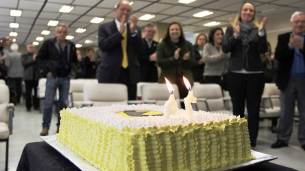A imagem mostra um bolo de aniversário em primeiro plano. Ele tem velas formando o número 79 e o símbolo do Daer na cobertura. Ao fundo, público no auditório do Caff bate palmas durante o parabéns a você.
