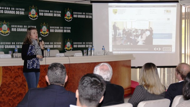 A foto mostra a relações públicas do Daer, Susane Goerck, falando ao microfone para o público presente no ato de aniversário do Daer. Ao fundo, no retroprojetor, a tela inicial do site novo do Daer.