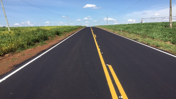 A foto mostra uma rodovia com asfalto e pintura renovados. O trecho, em reta, possui lavouras de grãos e postes de eletricidade às margens.