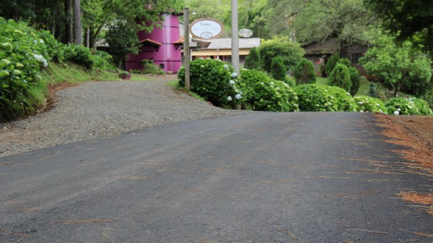 A foto mostra um trecho com asfalto recém implantado na estrada chamada Caminho das Pipas, em Rolante. Ao fundo, arbustos com hortências e uma pipa cor de rosa marcando a entrada para uma cantina colonial.