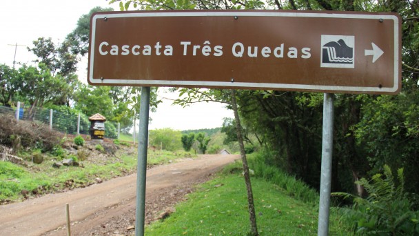 A foto destaca, em primeiro plano, uma placa indicando a localização da Cascata Três Quedas, em Rolante. Ao fundo, a estrada Caminho das Pipas, ainda sem pavimentação e, mais atrás, uma pipa de madeira indicando a entrada de uma cantina colonial.