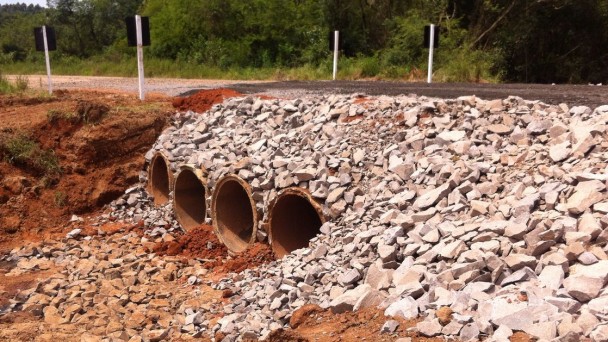 A foto mostra a rodovia ERS-130, não pavimentada. Em destaque, os quatro bueiros do trecho recém reconstruídos abaixo da pista e o aterro com pedras já recuperado.
