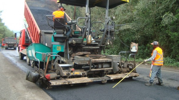 Na foto, dois operários trabalham no recapeamento de uma estrada. Um está operando na pista e o outro está dirigindo a máquina.