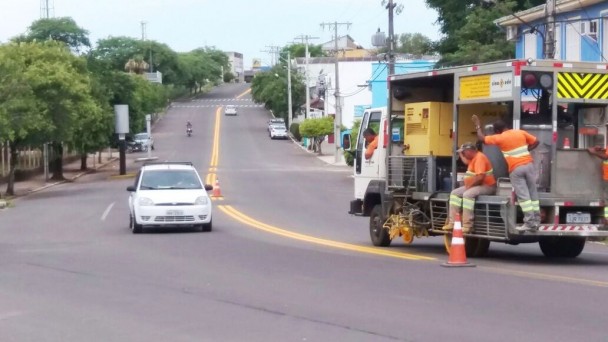 Na foto, uma máquina executa a pintura da faixa central da pista na ERS-409, no perímetro urbano de Vera Cruz. Há funcionários sentados no veículo. A pista tem cones de sinalização e há carros e motos passando no sentido contrário.