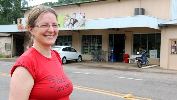 Na foto, a comerciante Maria Jeni Feltes aparece em primeiro plano, com a camiseta do supermercado dela. O estabelecimento aparece logo atrás, às margens da rodovia VRS-483, recém asfaltada.