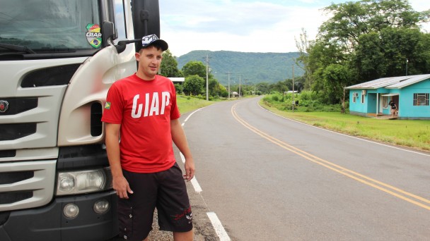 Na foto, o caminhoneiro Adriano Kaspary aparece em primeiro plano, logo em frente ao caminhão, estacionado no acostamento da VRS-843, em Feliz. Ao lado, aparece a rodovia pavimentada e com as faixas pintadas. Às margens, uma casa azul e árvores.
