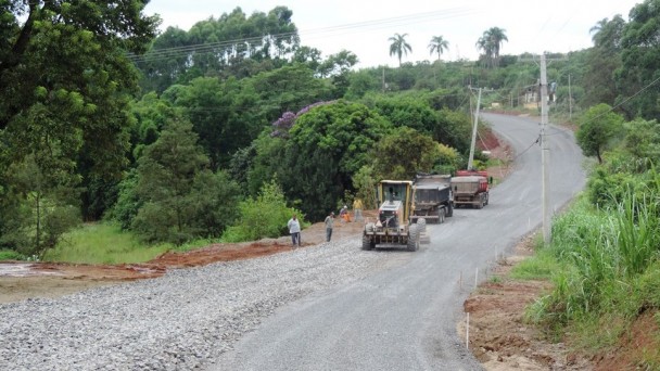 Na foto, um trator e dois caminhões percorrem o trecho em obras da VRS-874. A rodovia está com a base asfáltica implantada, boa parte dela com rachão.