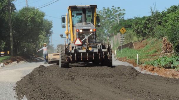 Na foto, um trator passa por cima do material asfáltico usado para compor a base das obras de pavimentação da VRS-874. Um operário orienta os trabalhos, ao fundo.