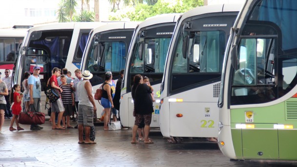 A foto mostra sete ônibus estacionados no setor de embarque da rodoviária de Porto Alegre, lado a lado. Passageiros com bagagens fazem fila à frente de um deles.