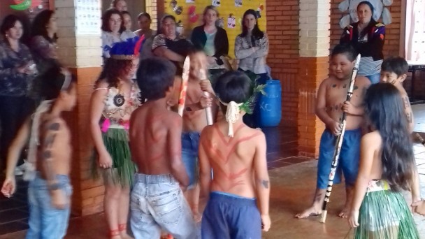 Na foto, um grupo de crianças indígenas, vestidas e pintadas conforme a cultura local, fazem uma apresentação no pátio de uma escola, assistidas por professores ao fundo.