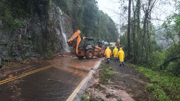 A foto mostra um grupo de operários com capas de chuva amarelas, junto a uma retroescavadeira, trabalhando para desbloquear a rodovia BRS-470, na Serra, que está obstruída por queda de barreiras