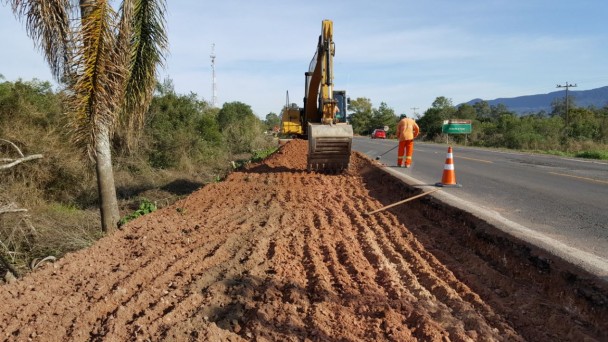 A foto mostra uma retroescavadeira trabalhando na escavação de uma das faixas da RSC-287, em Santa Maria. Um operário dá assistência ao trabalho.