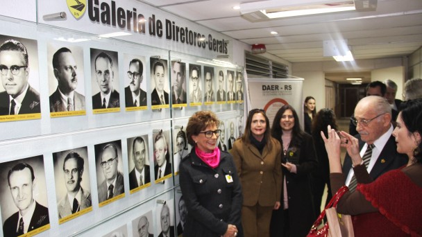 A foto mostra a recém inaugurada Galeria dos Ex-Diretores do Daer, com pessoas posando para fotos na frente do painel de vidro que reúne as fotos dos dirigentes.